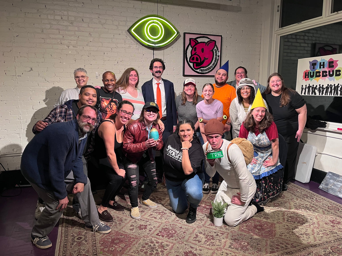 A group of 15 people posing together in a room with a white brick wall, wearing costumes and hats, with a neon eye sign and a pink pig picture on the wall behind them.